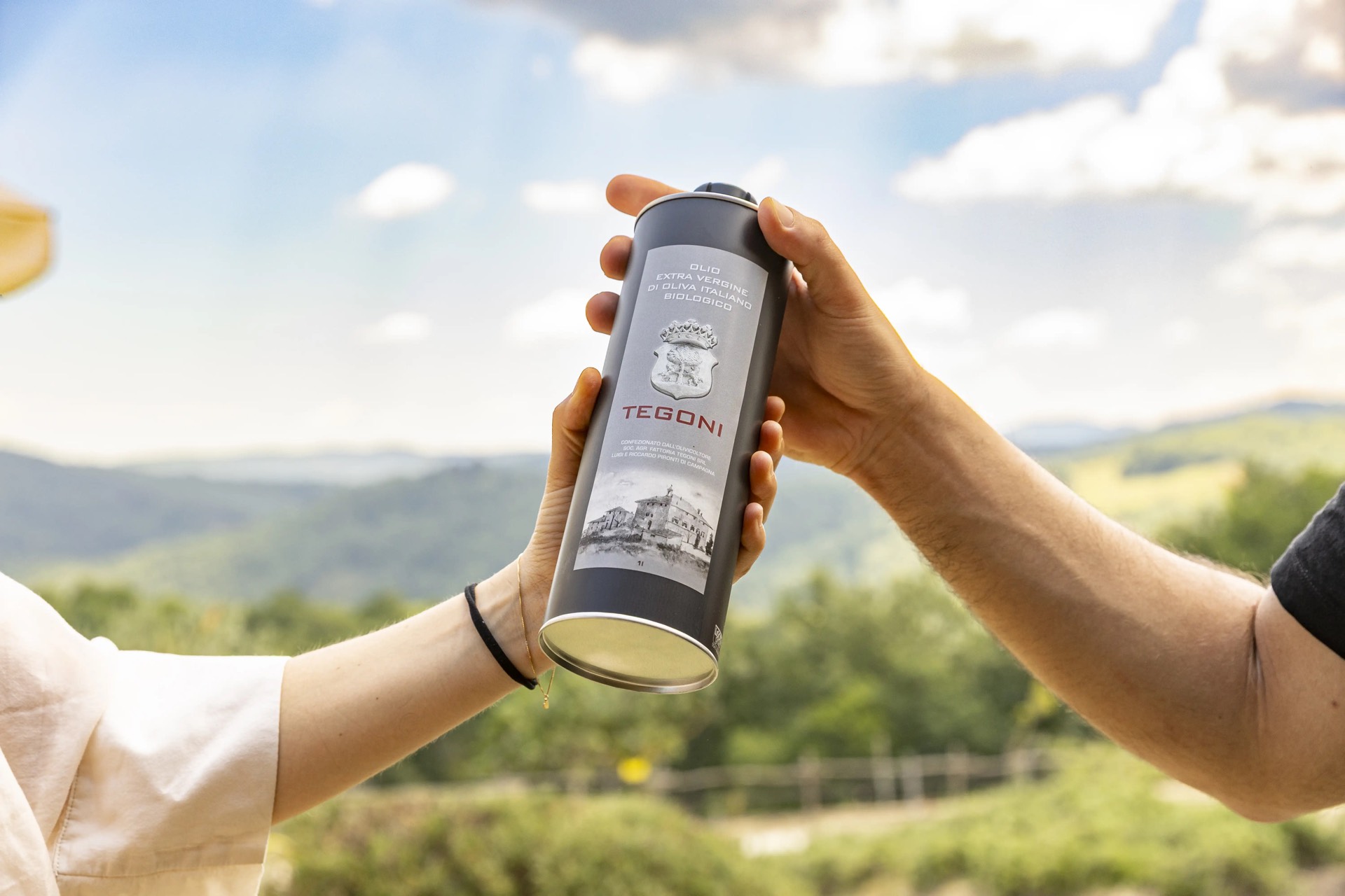 Le mani porgono la latta dell'olio extravergine Tègoni, colline sullo sfondo